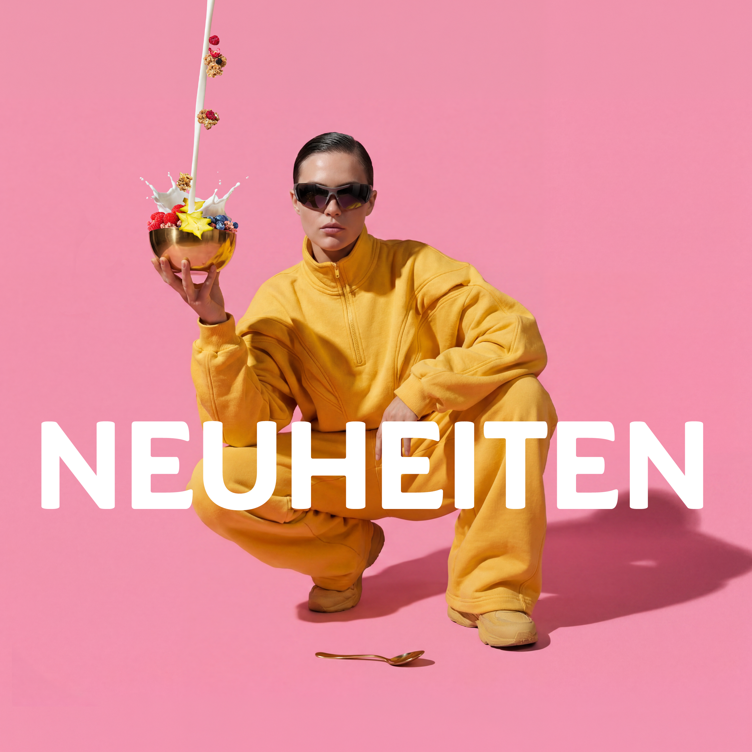 Dunkelhaarige Frau mit Sonnenbrille und gelbem Trainingsanzug, die vor rosafarbenem Hintergrund in der Hocke kniet. Sie schaut ernst in die Kamera und hält mit der rechten Hand eine goldene Schale mit Bio-Knuspermüsli, Sternenfrüchten sowie Himbeeren und Heidelbeeren. Von oben fließt ein Milchstrahl in die Schüssel, außen rum fliegen Cluster und Beeren. Unter der Frau liegt ein goldener Löffel. In der Mitte des Bilder ist der Schriftzug "NEUHEITEN" zu sehen.
