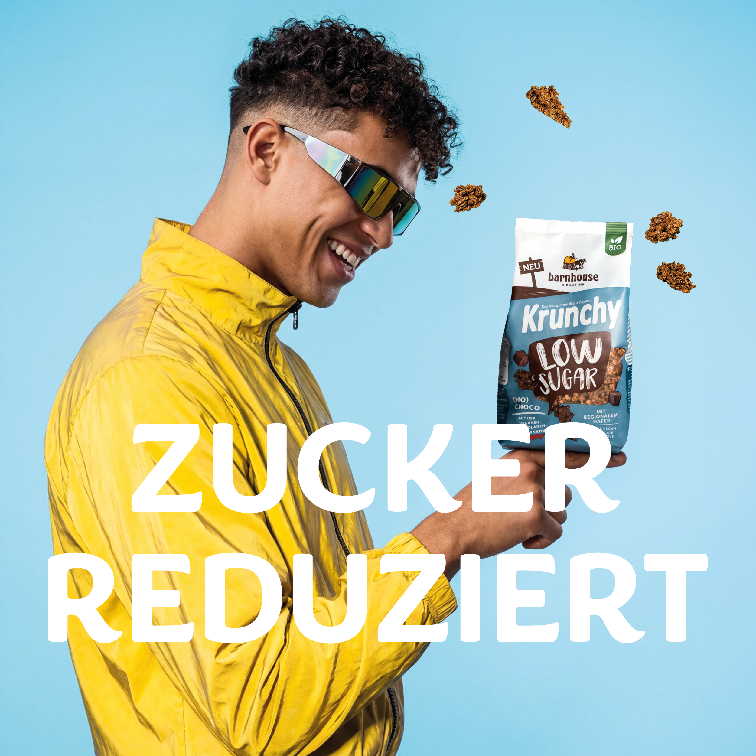 Junger Mann mit dunklen Locken und Sonnenbrille in gelber Trainingsjacke. Auf seinem ausgestreckten Finger steht ein braun-blauer Beutel Krunchy Low Sugar (No) Choco, den er glücklich angrinst. Um den Beutel herum fliegen Cluster. In der Mitte des Bilder ist der Schriftzug "ZUCKERREDUZIERT" zu sehen.
