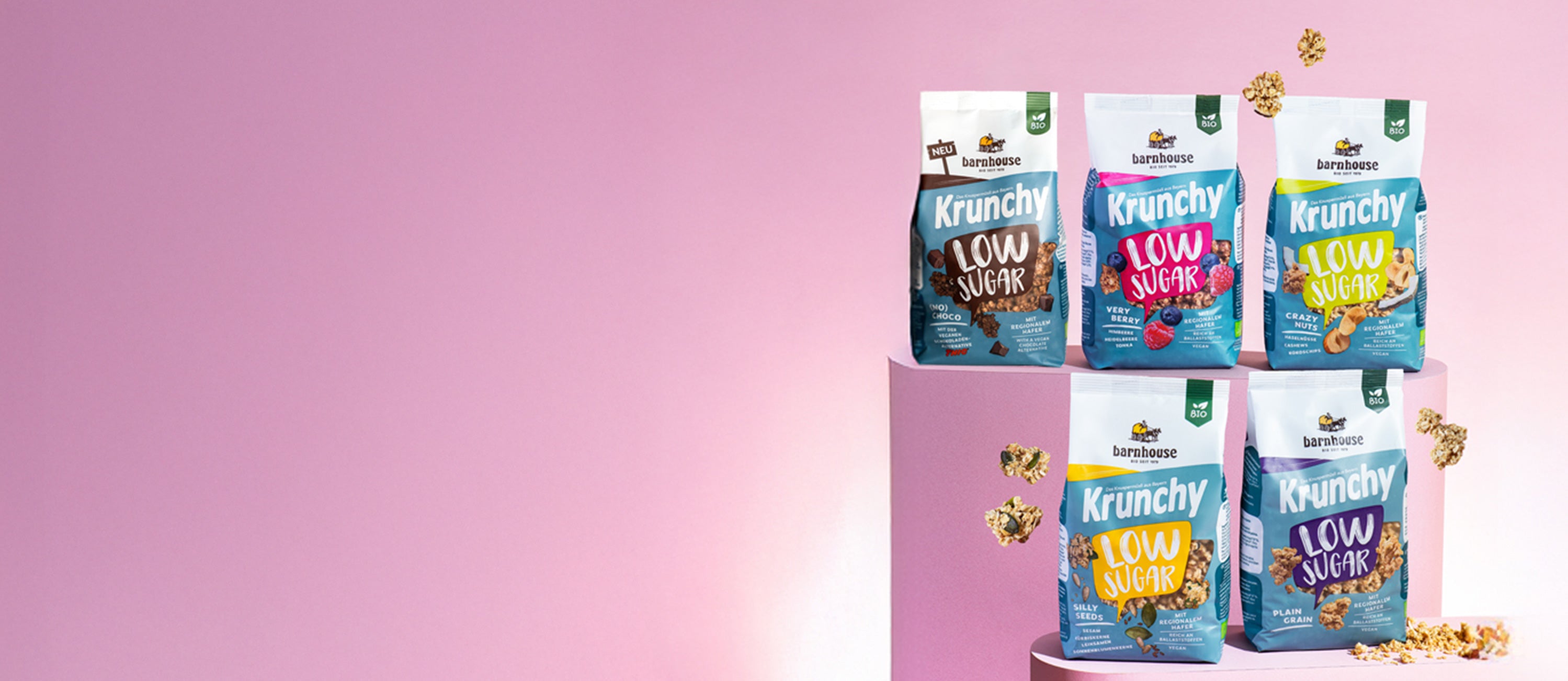 Blick von vorne auf die fünf Low Sugar Krunchys Plain Grain, Crazy Nuts, Very Berry (No) Choco und Silly Seeds, die auf zwei verschiedenen rosa Sockeln stehen. Außenrum fliegen Cluster.