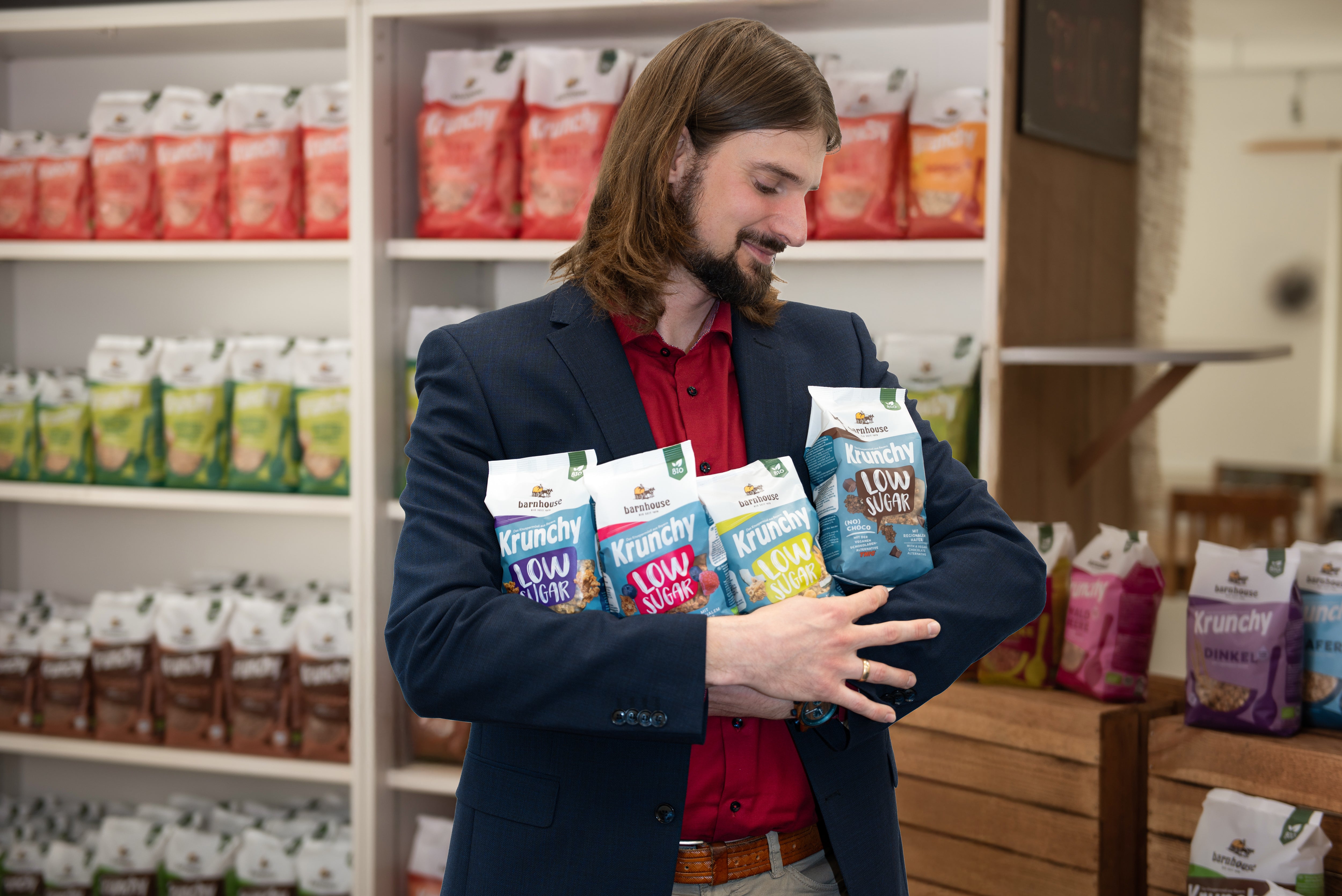 Barnhouse Personalleiter Stephan von Ahnen mit Hemd und Sakko gekleidet. Er steht vor einem mit Krunchy befüllten Regal und hält vier Sorten Krunchy Low Sugar im Arm.