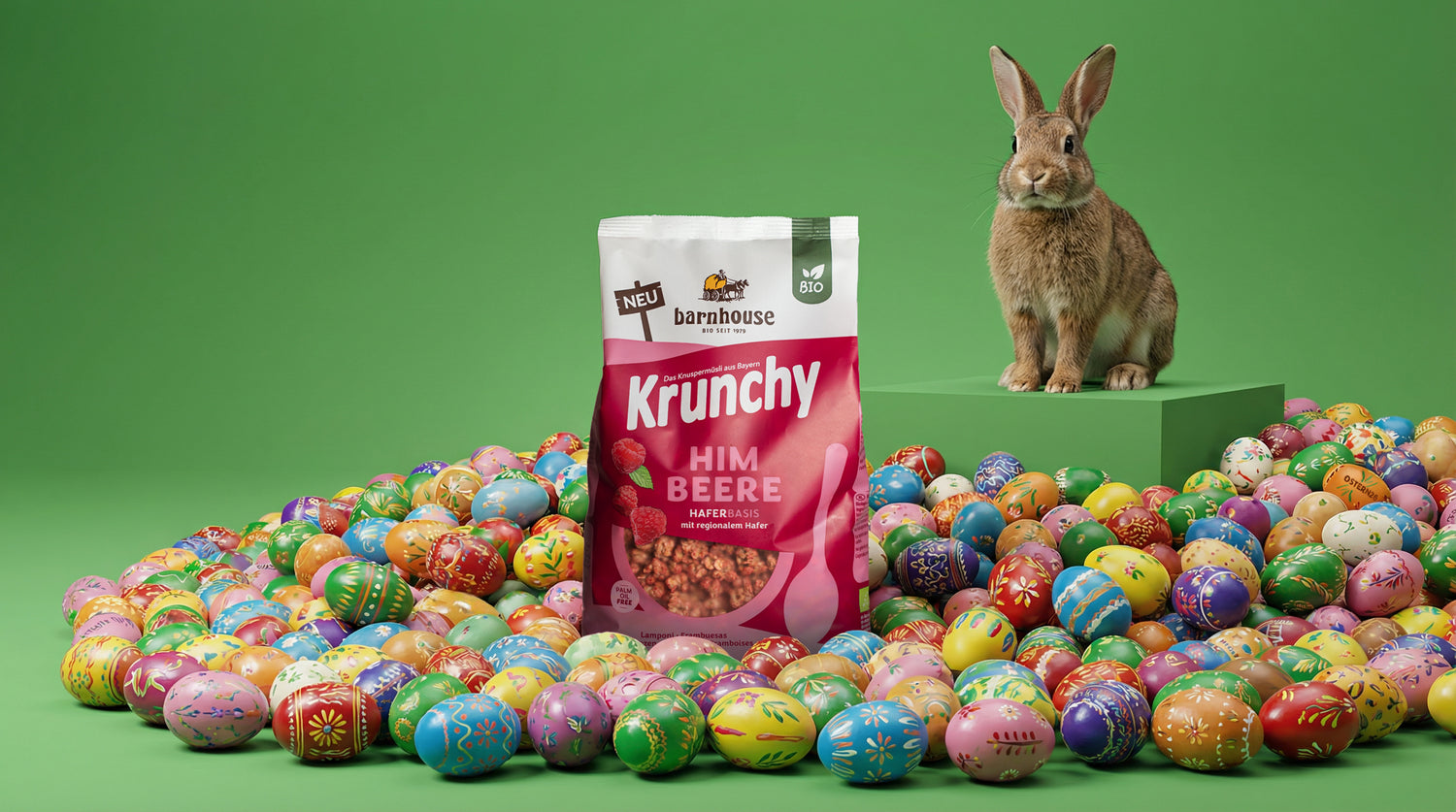 Brauner Hase, der auf einem viereckigen, grünen Podest sitzt. Am Boden liegen jede Menge bunte Ostereier. Mittendrin steht ein Beutel Krunchy Himbeere.