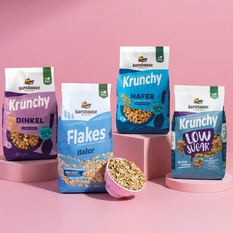 Blick von vorne auf die vier Basis-Produkte Dinkel 30% weniger Zucker, Flakes Hafer, Hafer 30% weniger Zucker sowie Low Sugar Plain Grain von Bio-Knuspermüsli-Hersteller Barnhouse, die auf verschiedenen, rosafarbenen Sockeln stehen. An dem Beutel Flakes Hafer lehnt eine rosa Schale mit den Flakes.