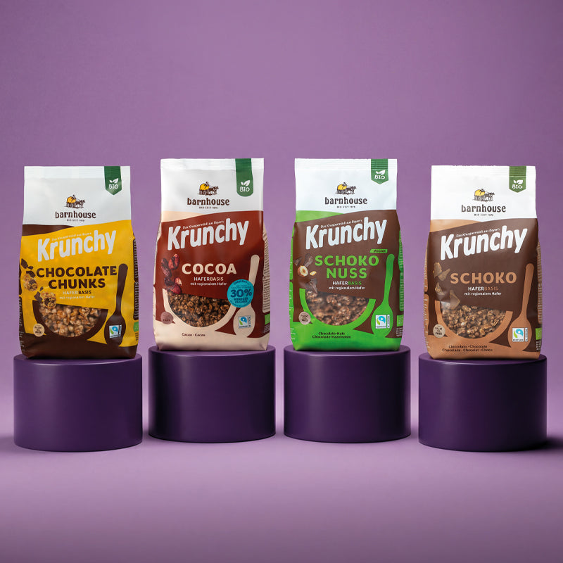 Blick von vorne auf die vier schokoladigen Krunchys Chocolate Chunks, Cocoa weniger Zucker, Schoko-Nuss und Schoko von Bio-Knuspermüsli-Hersteller Barnhouse, die allesamt auf einem eigenen, violetten, runden Sockel stehen. Der Hintergrund ist in einem helleren Lila.