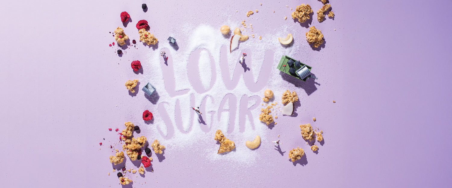weißer Schriftzug "Low Sugar" auf violettem Hintergrund. Außenrum liegen jede Menge Müsli-Cluster, Beeren, Nüsse und Kokosstücke. Männchen-Bauarbeiter bearbeiten den Schriftzug mit verschiedenen Werkzeugen und Maschinen.