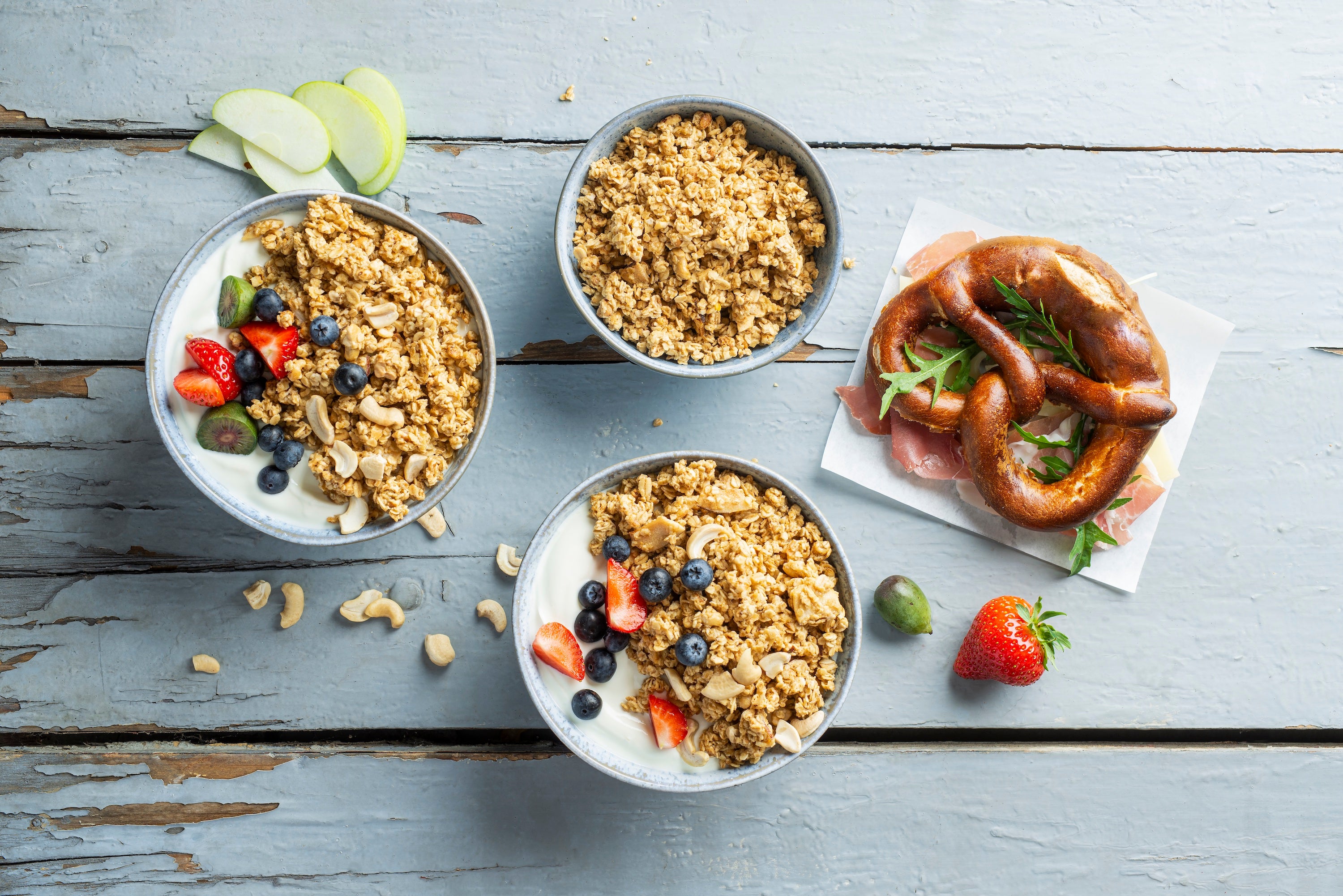 Oben steht eine blaue Müslischüssel mit Krunchy Low Sugar Crazy Nuts von Bio-Knuspermüsli-Hersteller Barnhouse. Im unteren Bereich steht eine weitere blaue Schale mit dem zuckerarmen Nuss-Müsli, allerdings mit Joghurt und Erdbeeren sowie Heidelbeeren als Topping. Links steht noch eine blaue Müslischale, die zudem noch mit zwei halben Minikiwis dekoriert ist. Rechts liegt eine Breze mit Schinken und Rucola auf einer weißen Serviette.